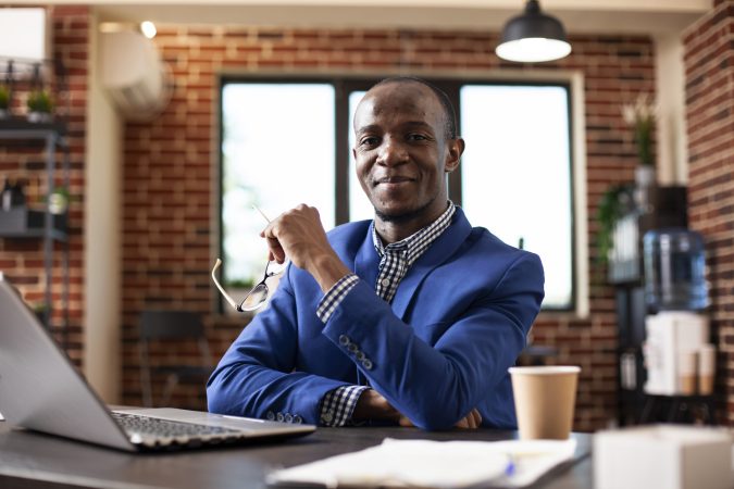 Portrait of businessman with spectacles in hand, sitting at desk and smiling at camera. African american entrepreneur in formal clothing posing at table in brick wall office, portraying confidence.