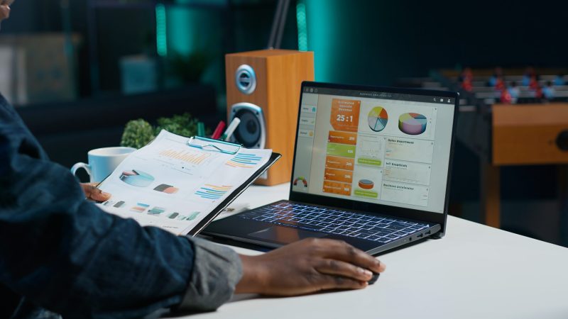 Woman sitting at home office desk, looking at business documents. African american freelancer in living room using notebook while reading financial charts on clipboard, camera A