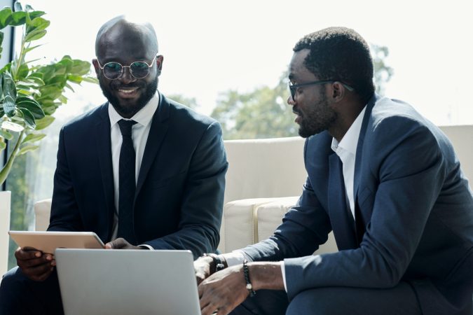 Young smiling African American businessman with tablet and his colleague having discussion of project points at meeting